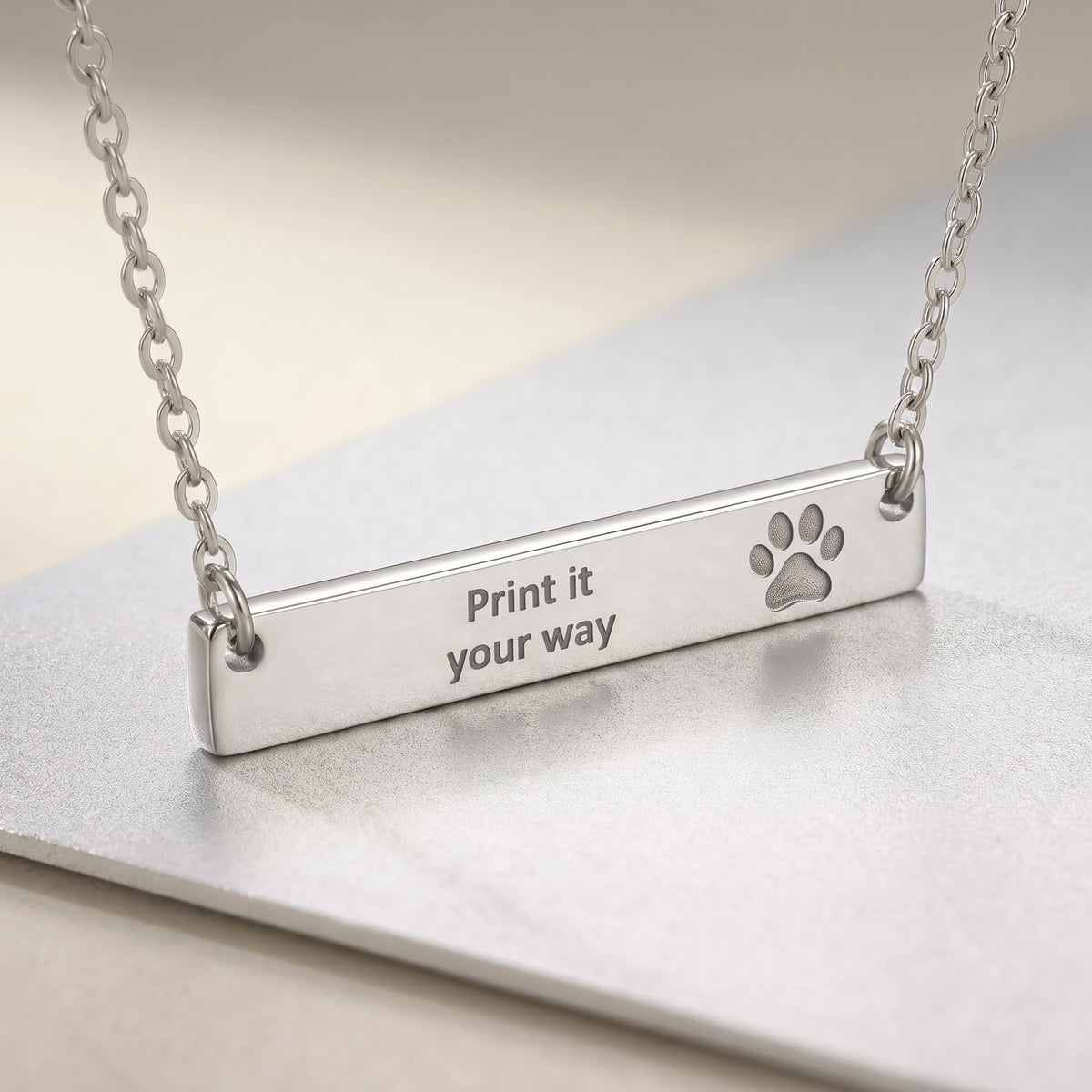 A silver bar necklace with a paw print and the engraved text 'Print it your way', resting on a metallic surface.
