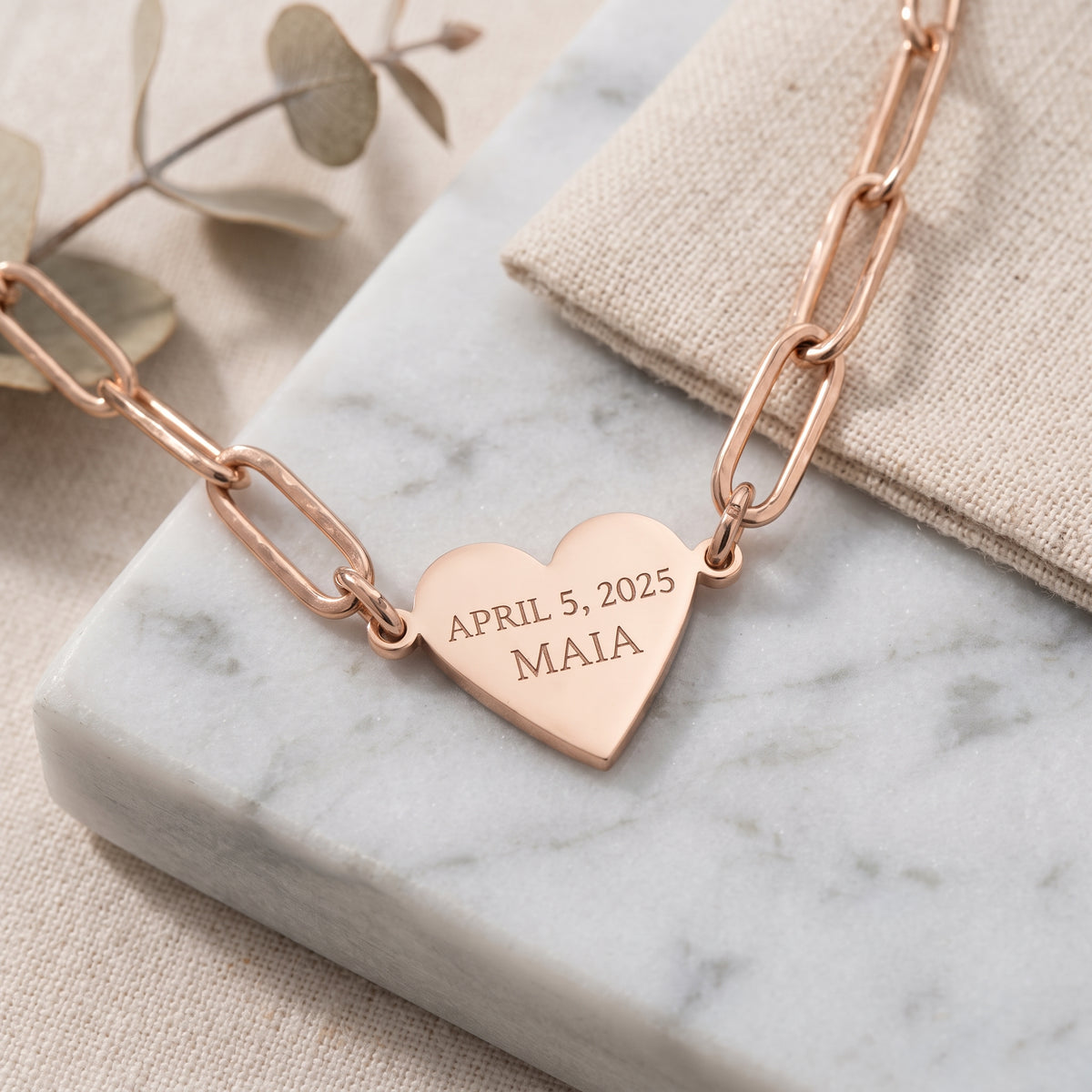 A rose gold heart-shaped pendant necklace engraved with the date 'APRIL 5, 2025' and the name 'MAIA', resting on a white marble slab with a beige linen cloth and dried eucalyptus leaves.